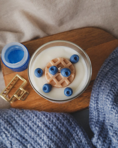 Vela em copo transparente com aroma de baunilha e mirtilos e com o topo da vela decorado com um waffle e vários mirtilos